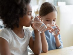 Two multi racial little girls sit at table in kitchen feels thirsty drink clean still natural or mineral water close up image. Healthy life habit of kids, health benefit dehydration prevention concept