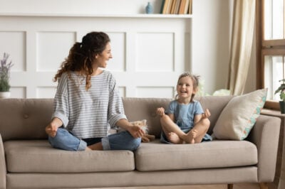Full length front view smiling young curly mother sitting on comfortable couch with cute playful little preschool daughter in lotus position. Happy mommy practicing yoga exercised with small child.