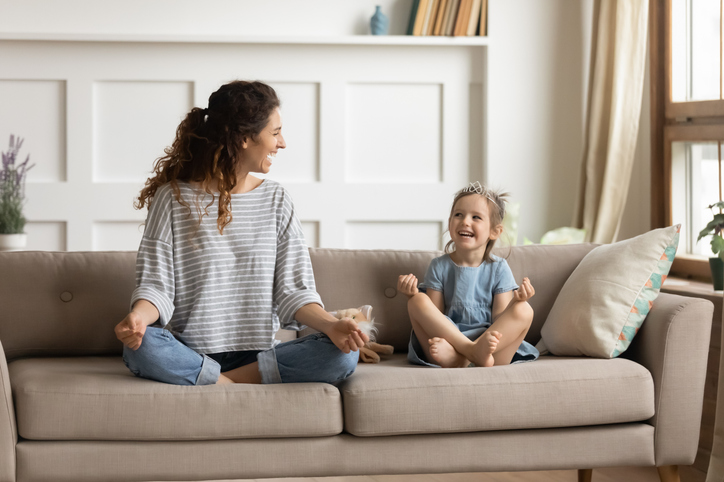 Full length front view smiling young curly mother sitting on comfortable couch with cute playful little preschool daughter in lotus position. Happy mommy practicing yoga exercised with small child.