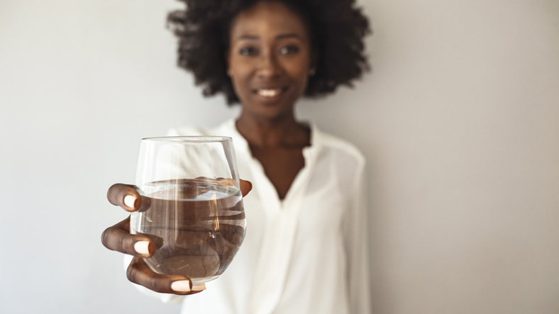Beautiful young woman smiling while holding a glass of water at home. Lifestyle concept. Happy young woman showing drinking glass with water at home. Hydration from a reverse osmosis system is her beauty secret