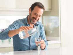 Middle age man drinking a glass of water with a happy face standing and smiling with a confident smile showing teeth