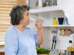 Senior woman drinking water while standing by window in kitchen background, people and healthy lifestyles