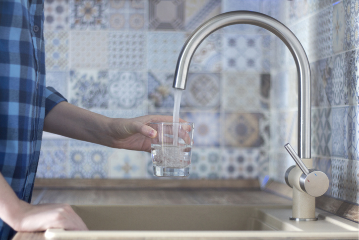 Woman in a blue plaid shirt pours water into the glass of the faucet in the kitchen
