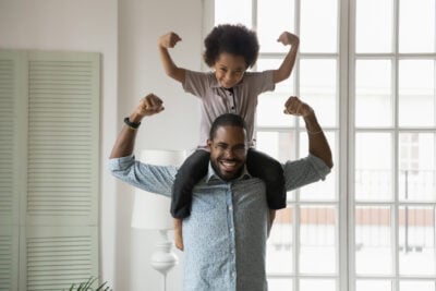 Small son sit on strong dad shoulders showing biceps. African family enjoy activity games at home, healthy fit lifestyle, two superheroes, vitamins for adults and children ad, happy Father Day concept