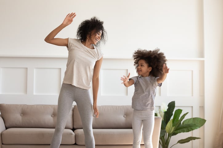 Happy smiling mother doing morning exercises with excited little preschooler daughter in stylish living room at home. Overjoyed black family dancing, jumping, having fun together.