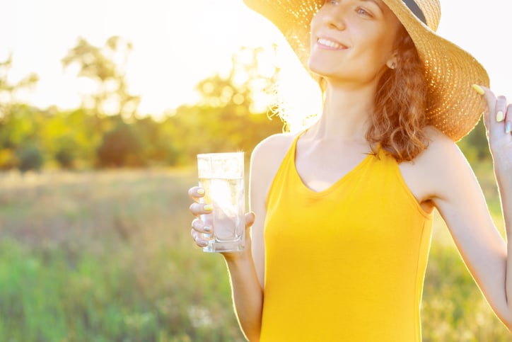 Happy beauty young woman female with pure glass of water wears yellow lite dress and summer hat smiling walking outdoor in green park at sunny warm light day. Active outdoor lifestyle leisure concept.