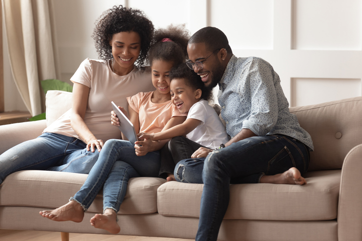Happy african family having fun with device at home, black parents and little children using digital tablet looking at screen sit on sofa together, cute kids hold computer laugh watch cartoons online