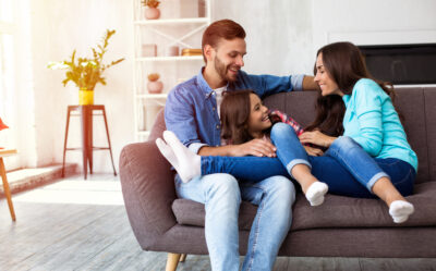 A beautiful family of father, daughter and mom are sitting on the sofa at home, laughing, hugging and playing with each other. Healthy indoor air quality with houseplants.