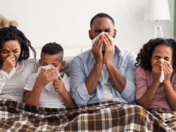 Portrait of sick young family of four people blowing runny noses while sitting together on the sofa with napkins and covered with blanket American parents and kids suffering from cold