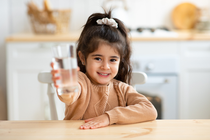 Healthy Drink. Cute Little Girl Holding Drinking Water In Kitchen, Adorable Female Child Sitting At Table And Giving Glass To Camera, Enjoying Refreshing Beverage, Closeup Shot With Selective Focus
