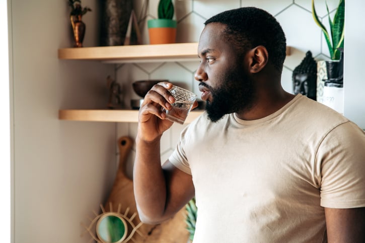 Healthy lifestyle. Handsome pensive healthy young African American bearded man, stands at kitchen, dressed in casual wear, drinks clean water from water filtration system, looks to the side