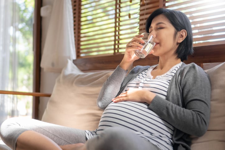 pregnant woman sitting on sofa drinking pure fresh glass of water in the morning. Happy healthy lifestyle.