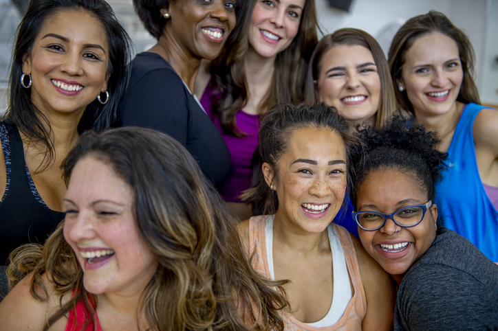 A large group of racially diverse middle aged woman, huddle together for a group portrait after a fitness class. They are each dressed comfortably in athletic wear and are smiling as they laugh with one another. womens health and wellness