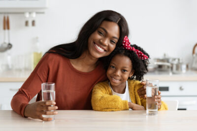 Happy African American Family Mother And Daughter Holding Glasses With Water While Sitting Together At Table In Kitchen, Smiling Black Mom And Female Child Enjoying Healthy Drink
