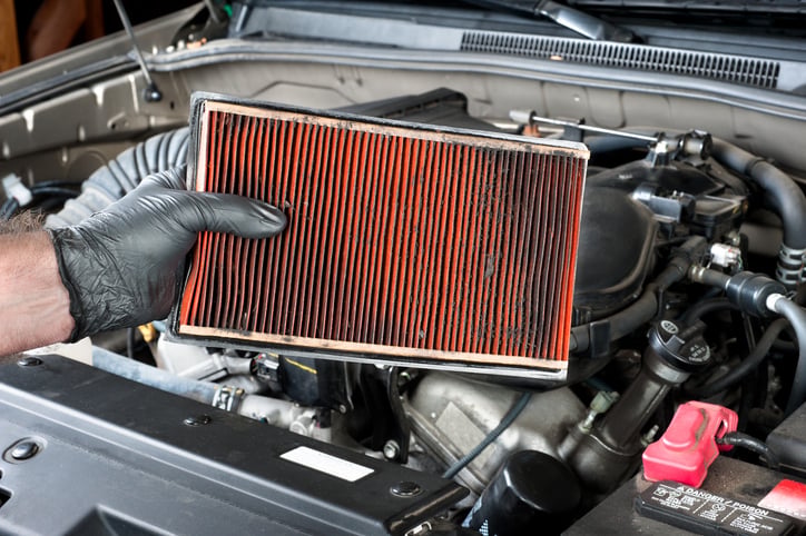 An auto mechanic wearing protective work gloves holds a dirty, clogged air filter for car over a car engine during general auto maintenance.